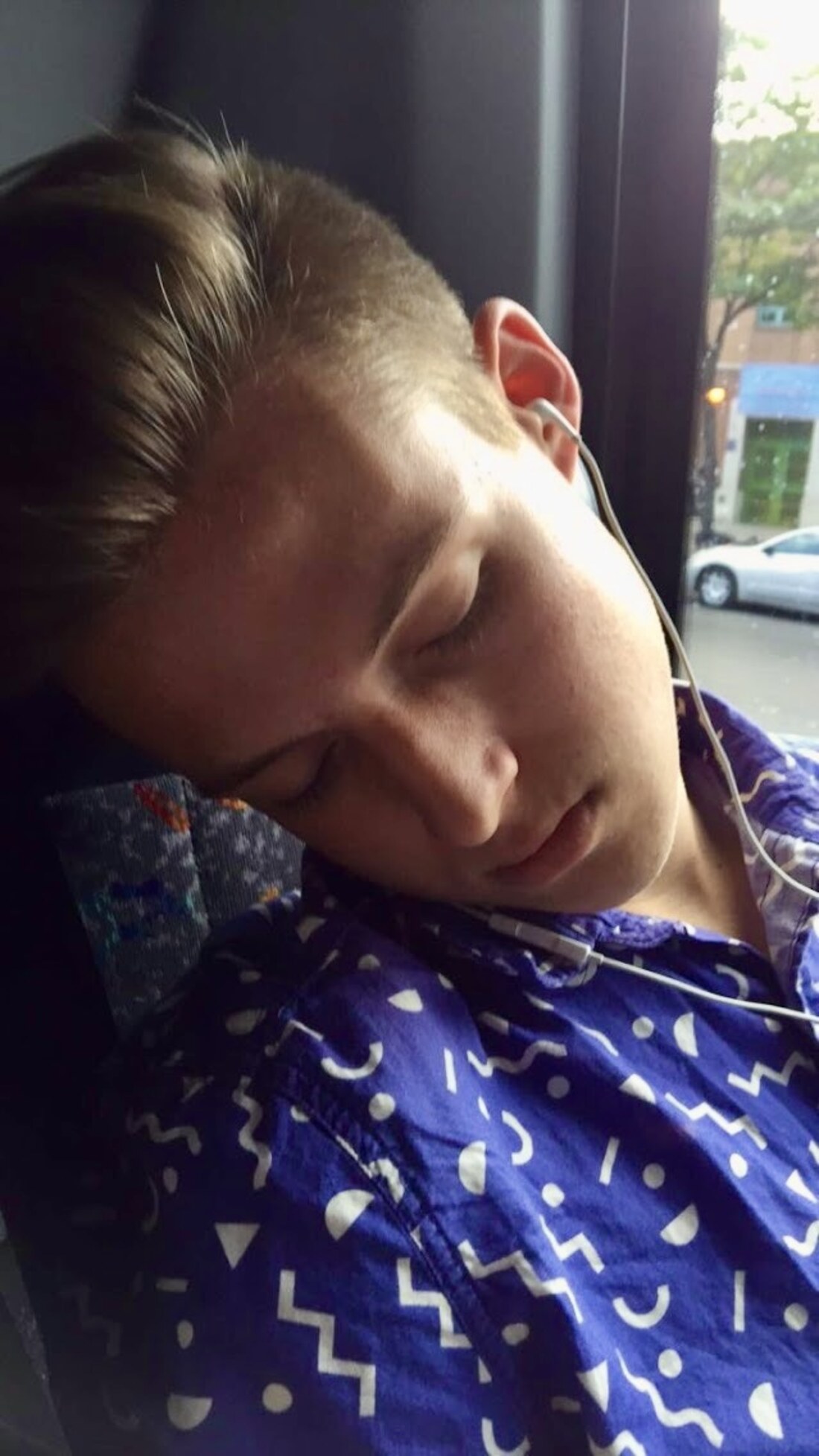 A teenager asleep against a bus window in a purple patterned shirt, wired earbuds still in