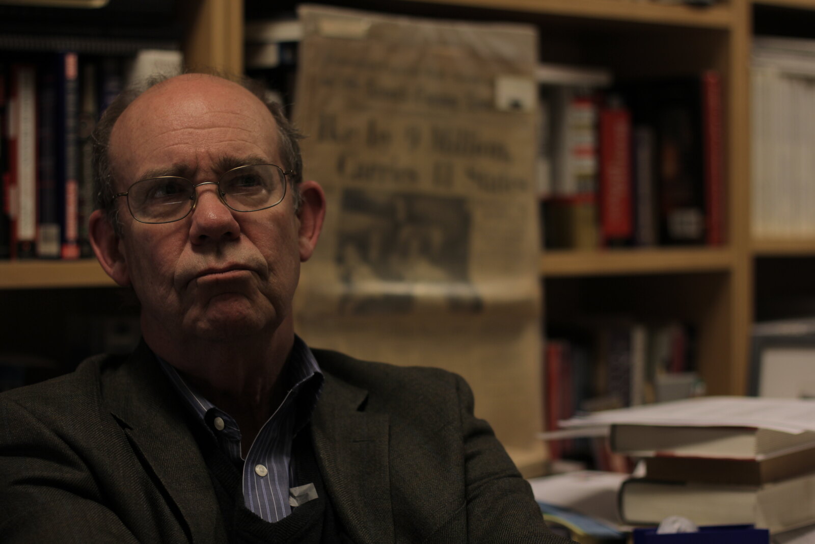 David Eisenhower mid-interview in his university office, bookshelves of presidential histories behind him and a framed 1956 newspaper on the shelf