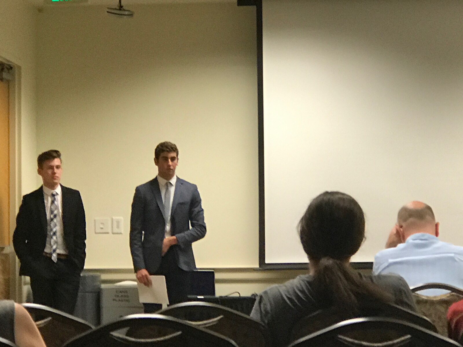 Two teenagers in suits standing at the front of a competition room beside a projection screen, facing a row of judges