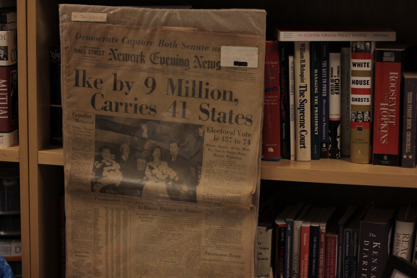 A close-up of the shelf: a preserved copy of the Newark Evening News, headline “Ike by 9 Million, Carries 41 States,” with a handwritten label reading Eisenhower