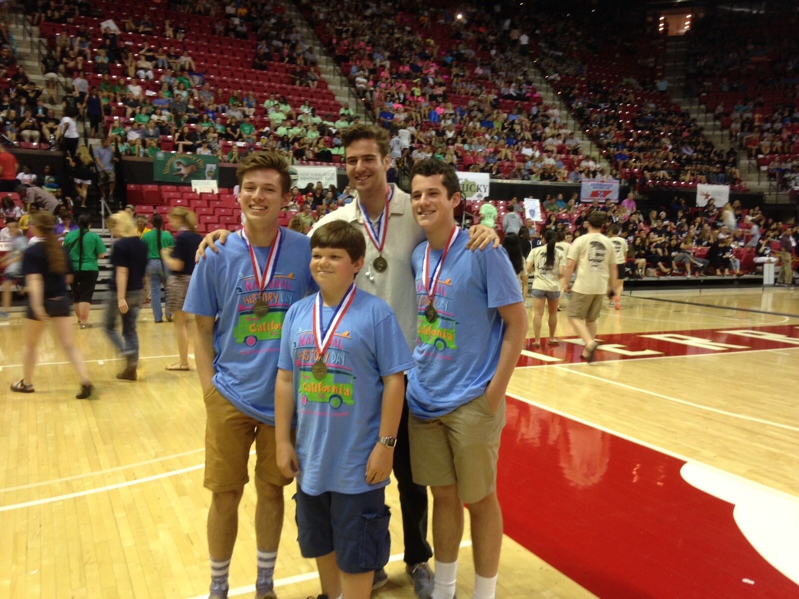 Four students in blue National History Day California shirts, wearing medals, standing on a basketball court in a packed arena