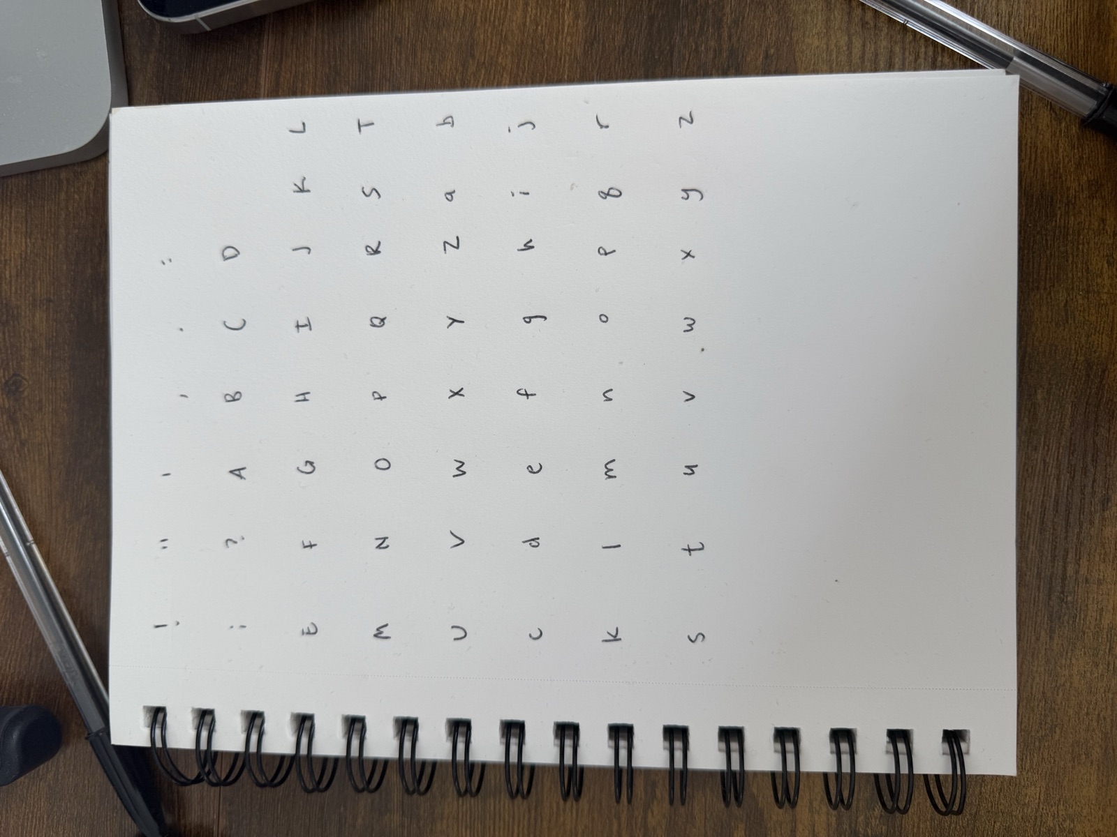 A spiral notebook page filled with a grid of hand-drawn letters, one glyph per cell