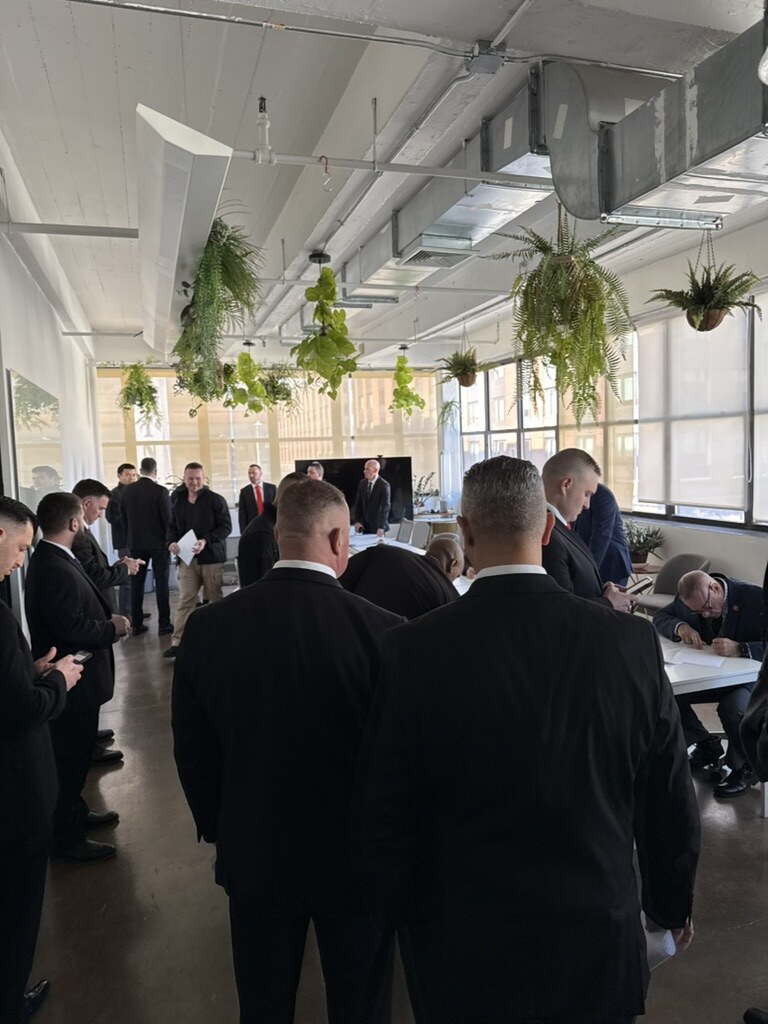 Protector agents in dark suits standing in a bright Brooklyn office with glass walls and hanging plants