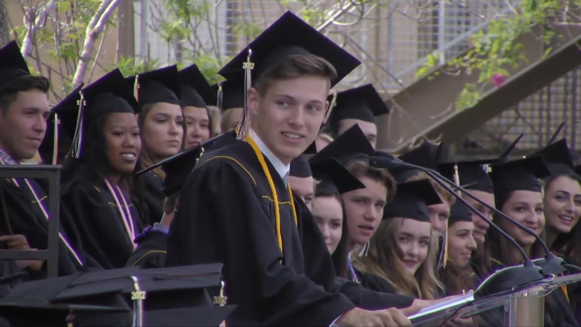 A graduate in cap and gown at the podium, seen from the side, looking up at the audience with a slight smile, rows of classmates in caps and gowns behind him