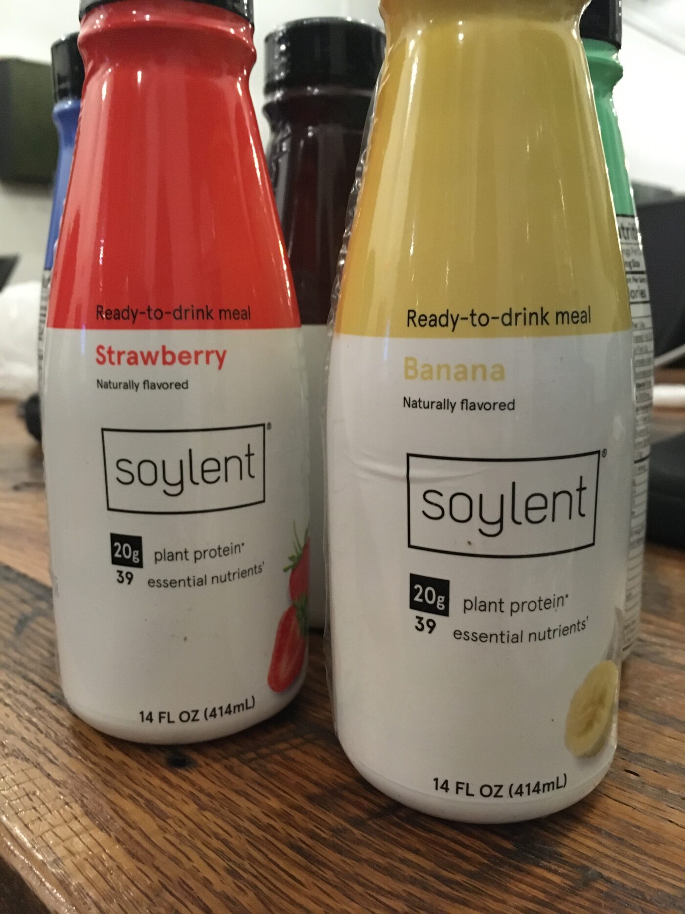 Two bottles of Soylent, strawberry and banana, with four more lined up behind them on a wooden table
