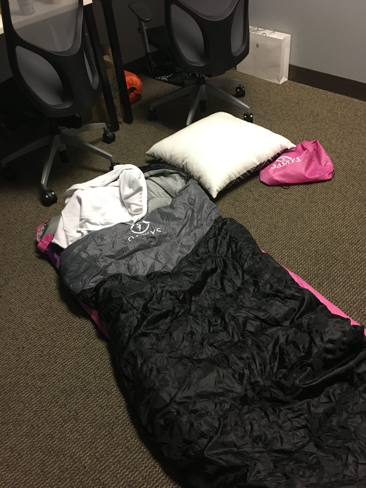 A sleeping bag, pillow and hoodie laid out on office carpet next to the wheels of two desk chairs